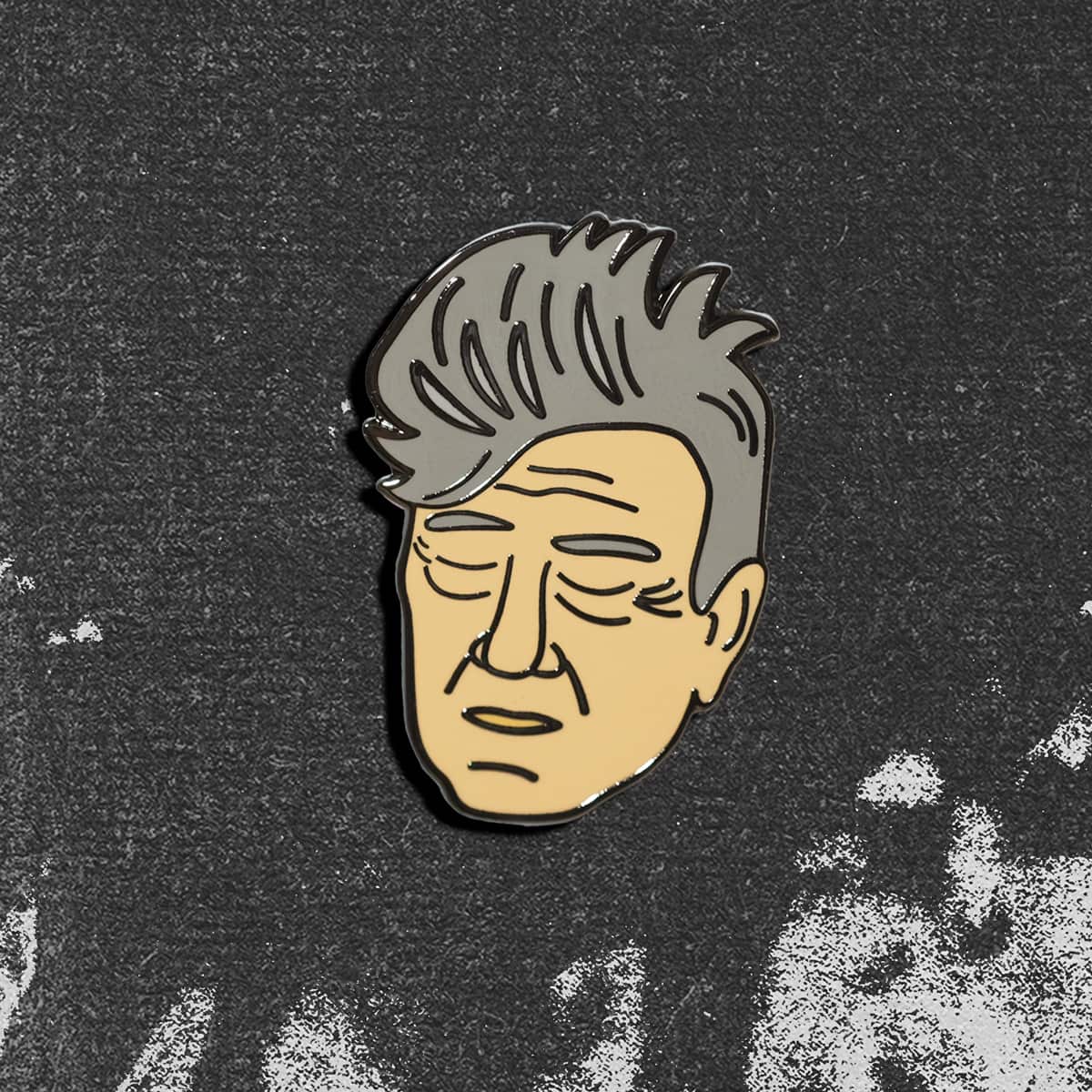 An enamel pin of David Lynch's face, shown on a dark textured background.