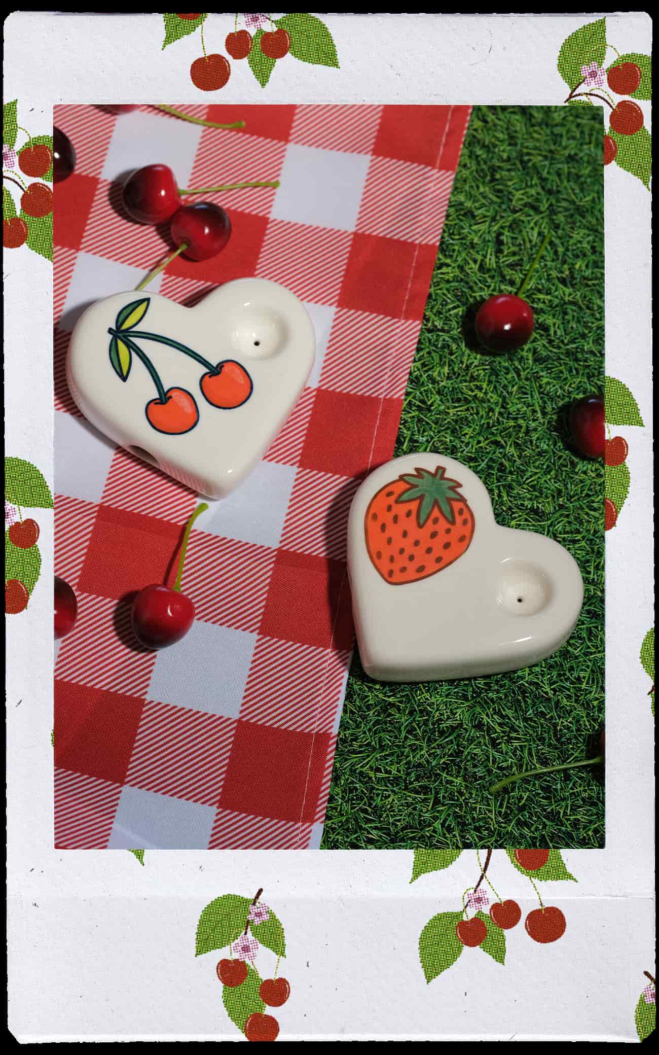 Two heart-shaped ceramic pipes with cherry and strawberry illustrations, styled on a red-and-white checkered blanket over artificial grass with scattered cherries. A white border with cherry illustrations frames the photo.