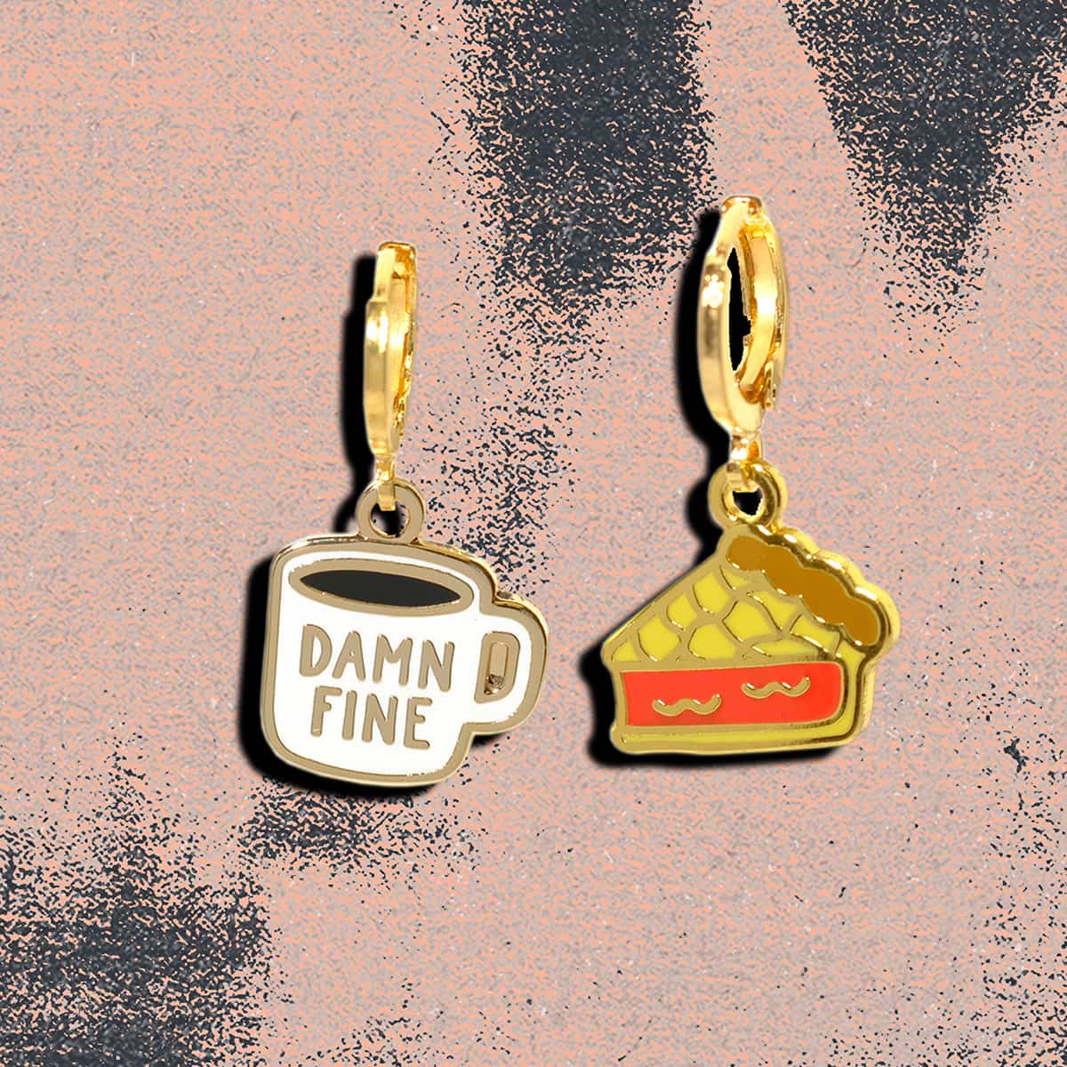 Two gold-tone enamel charms on a textured pink background. One charm is a white coffee mug with the words 'DAMN FINE' in gold. The other is a cherry pie, this earring set is referencing Twin Peaks