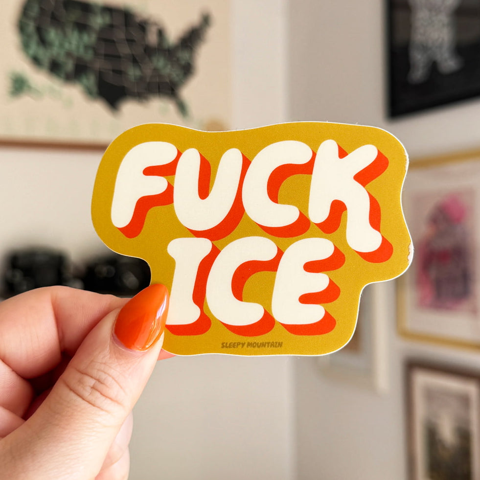 A hand with orange nail polish holds a green die-cut sticker that reads 'FUCK ICE' in bold white letters with red outlines, against a blurred indoor wall with framed pictures.