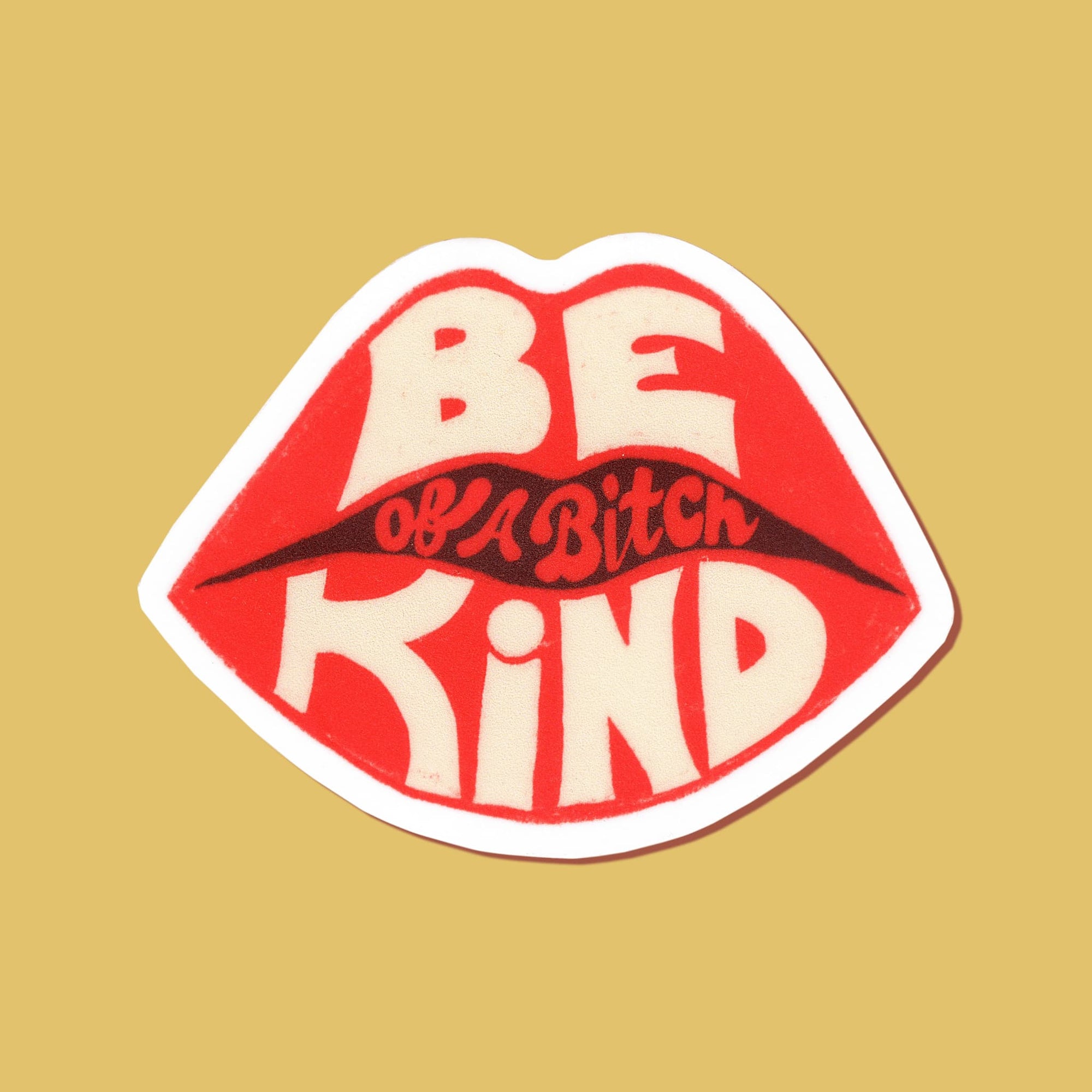 A die-cut sticker shaped like lips with the text 'BE KIND of a bitch' in red and cream lettering, shown on a solid mustard yellow background.