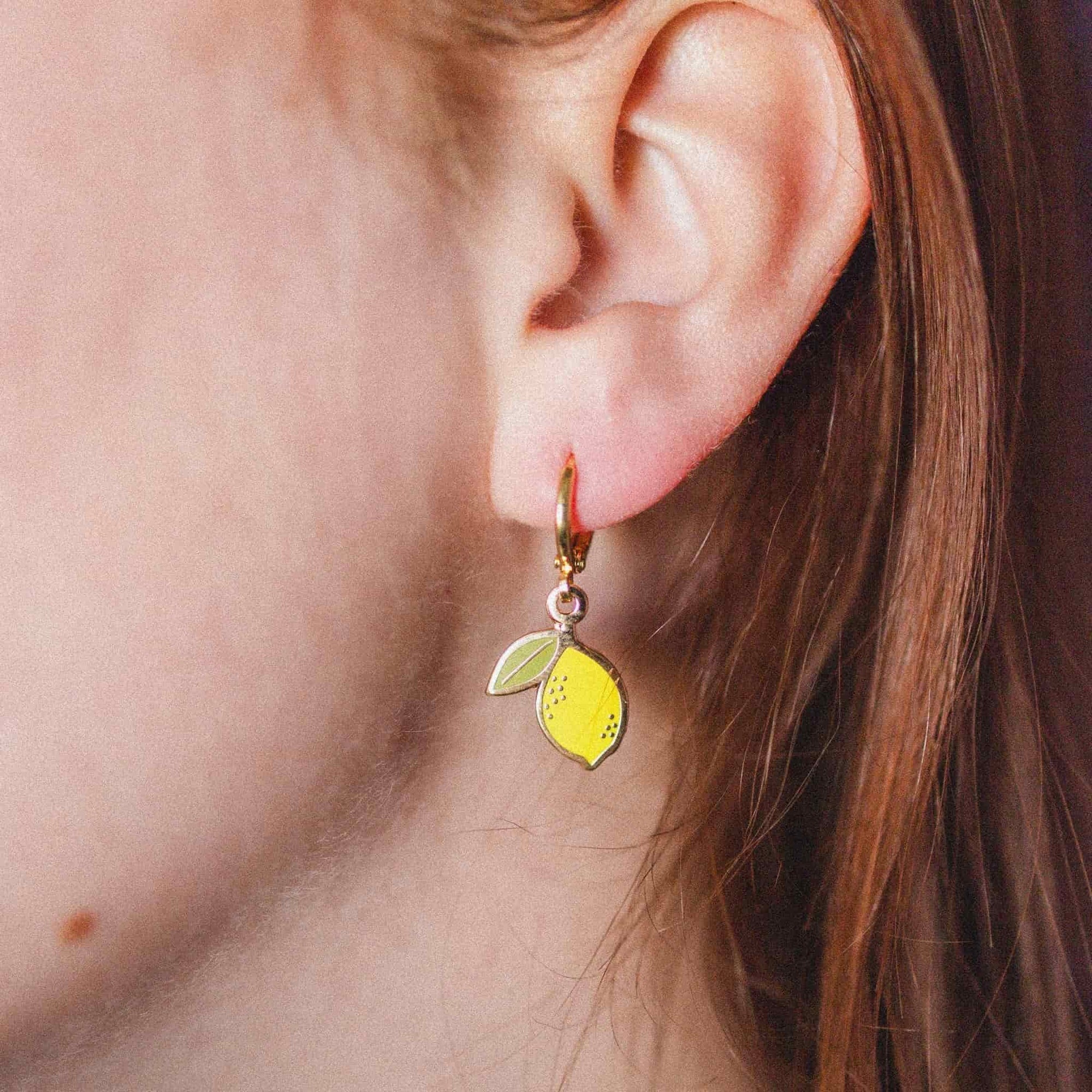 A close-up of a person's ear wearing a small dangling earring shaped like a lemon with a gold hoop and yellow-green enamel details.