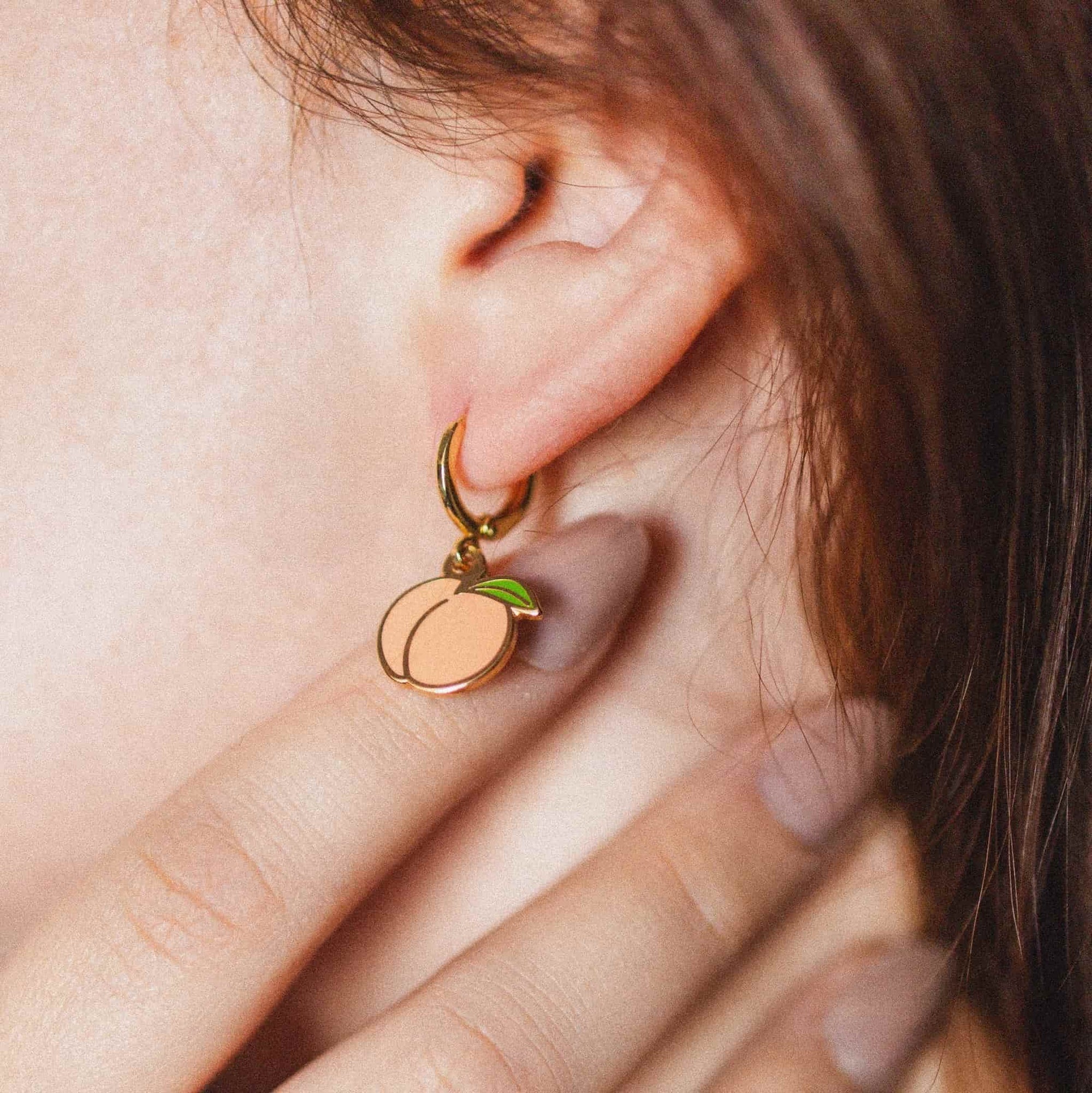 A close-up of a person's ear wearing a small gold hoop earring with a peach-shaped charm and a green leaf detail.