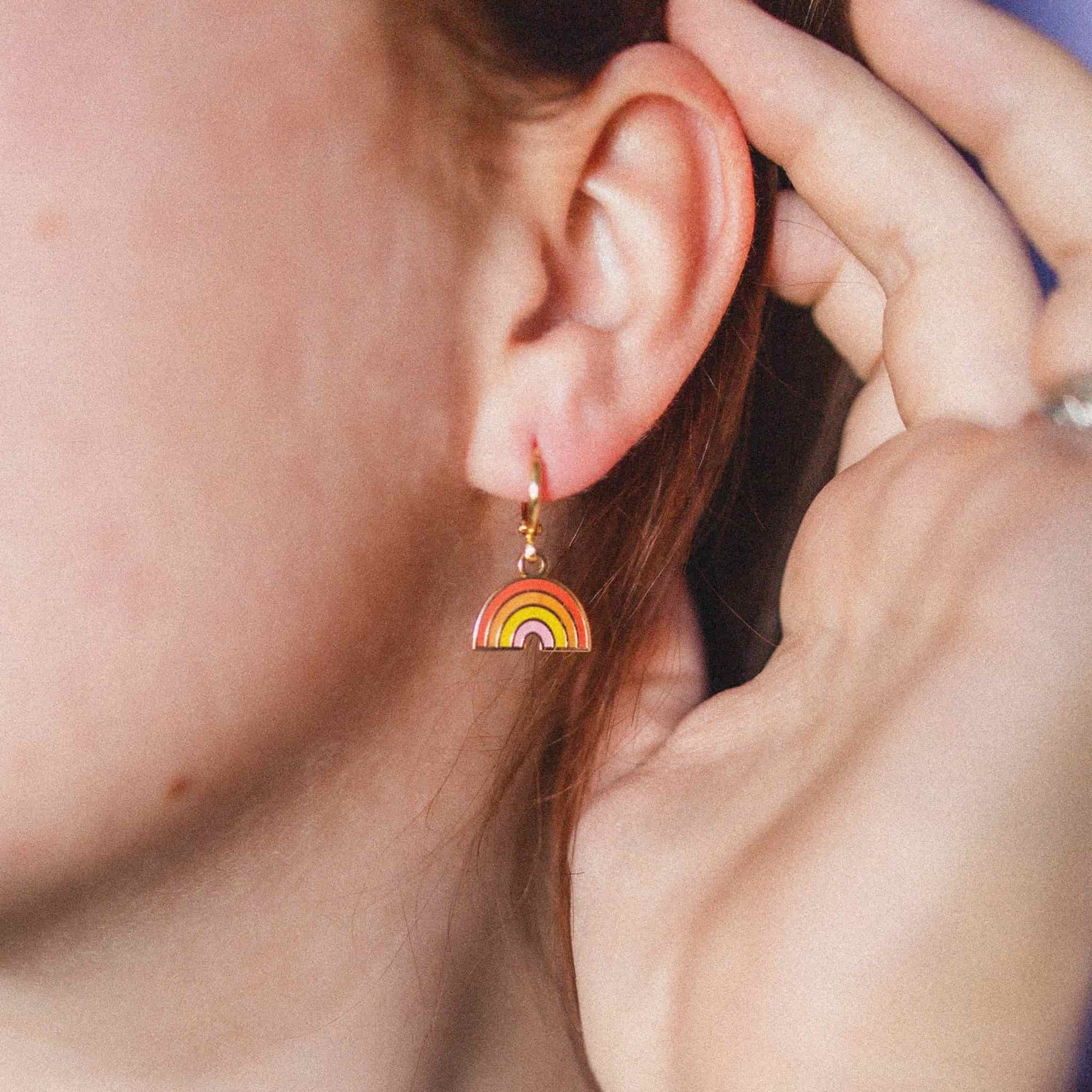 A close-up of a person's ear wearing a small rainbow-shaped dangle earring with gold-tone metal and red, orange, yellow, green, and purple enamel bands.