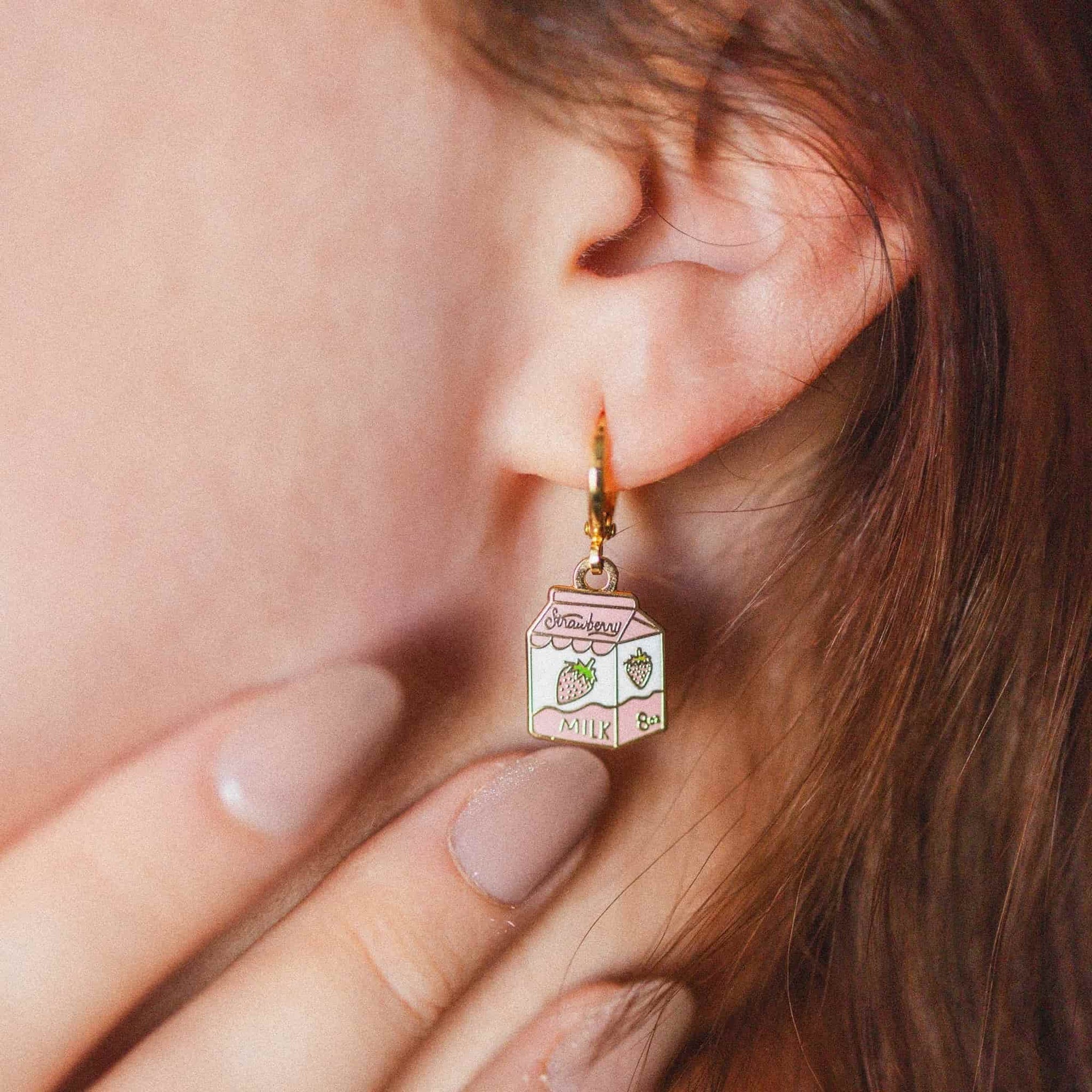 A close-up lifestyle shot of a person wearing a gold hoop earring with a small white milk carton charm featuring strawberries and the text 'Strawberry Milk'.